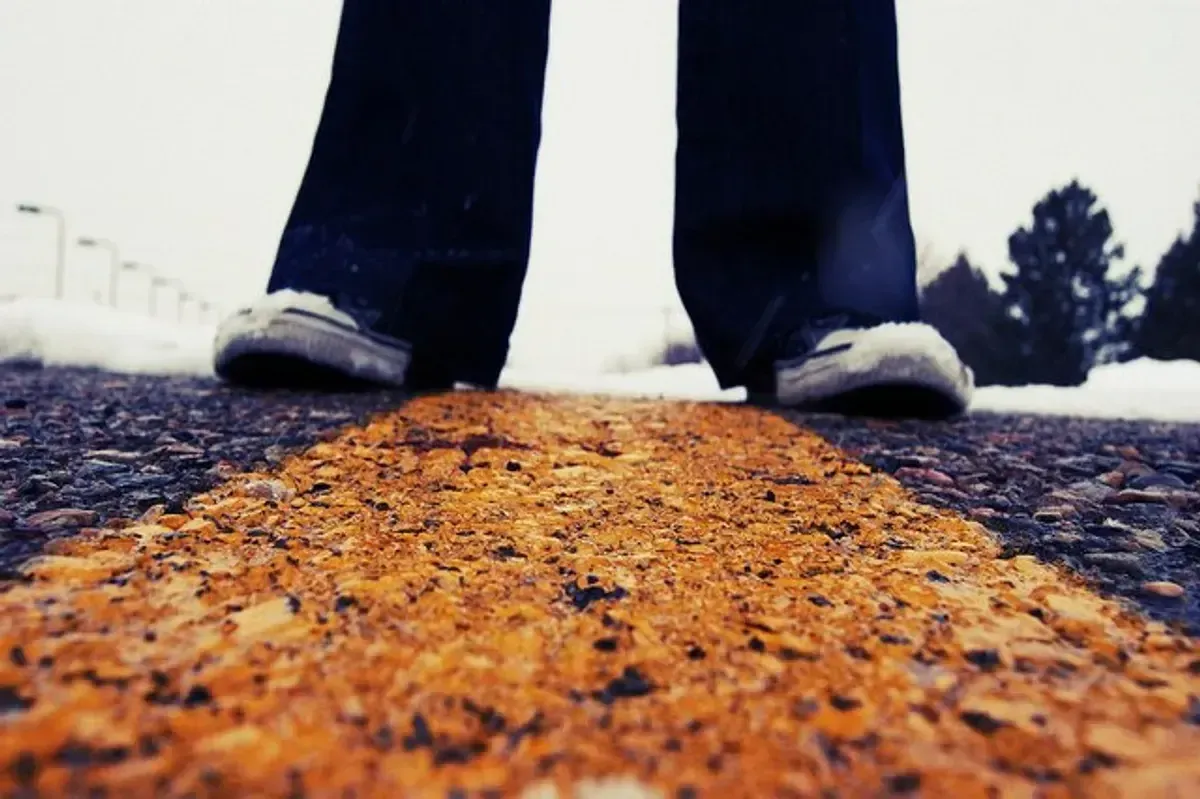 Primo piano di gambe di una persona in piedi in mezzo alla strada. L'immagine vuole evocare il trovarsi in due luoghi diversi, perché le gambe si trovano ognuna al di là della riga tracciata sulla strada