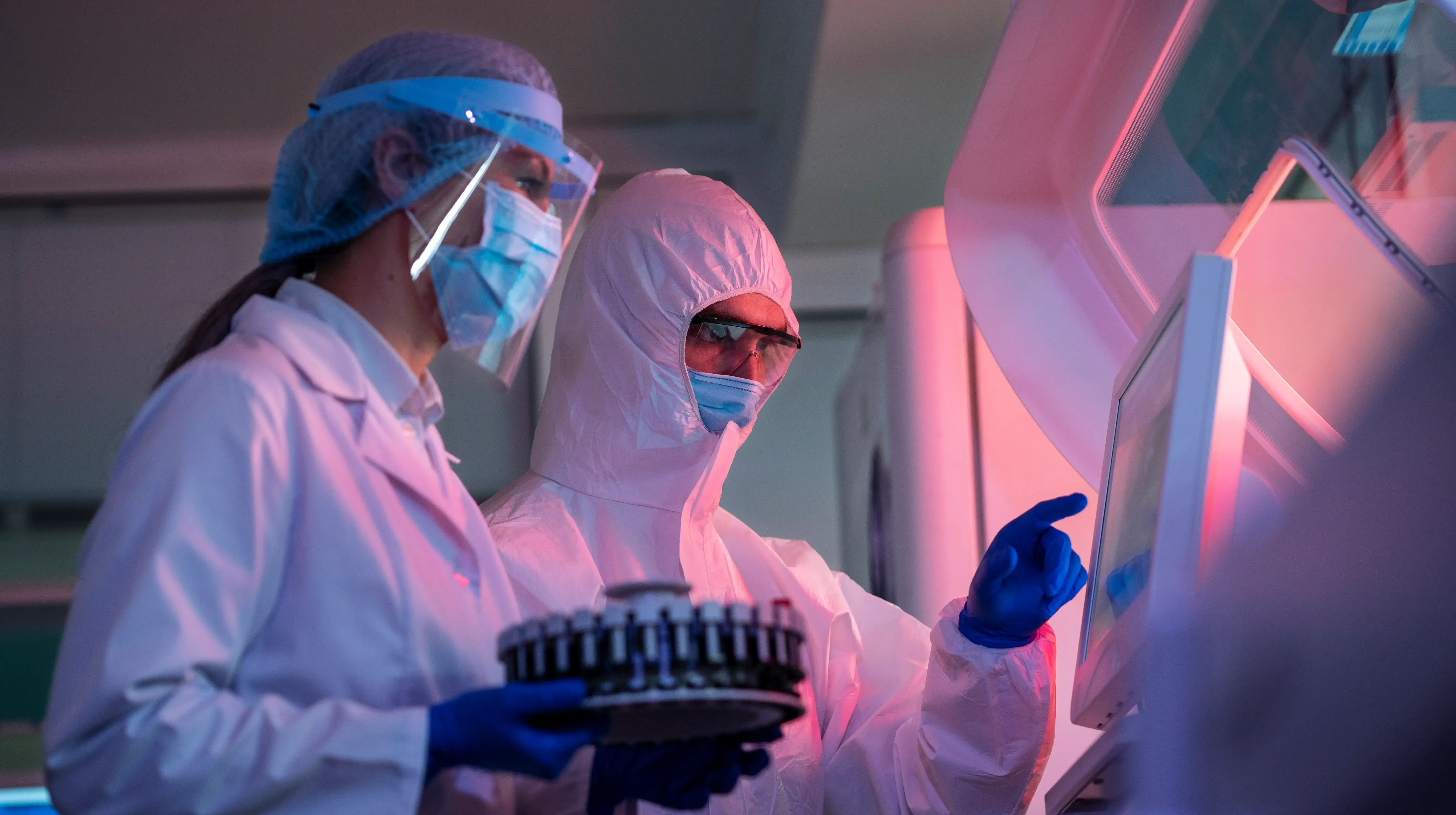 Foto di due ricercatori in un laboratorio con delle tute protettive e che lavorano con un macchinario