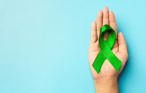 Immagine di una mano su sfondo celeste che tiene nel palmo una fiocco verde, simbolo della salute