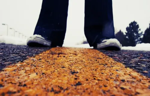 Primo piano di gambe di una persona in piedi in mezzo alla strada. L'immagine vuole evocare il trovarsi in due luoghi diversi, perché le gambe si trovano ognuna al di là della riga tracciata sulla strada