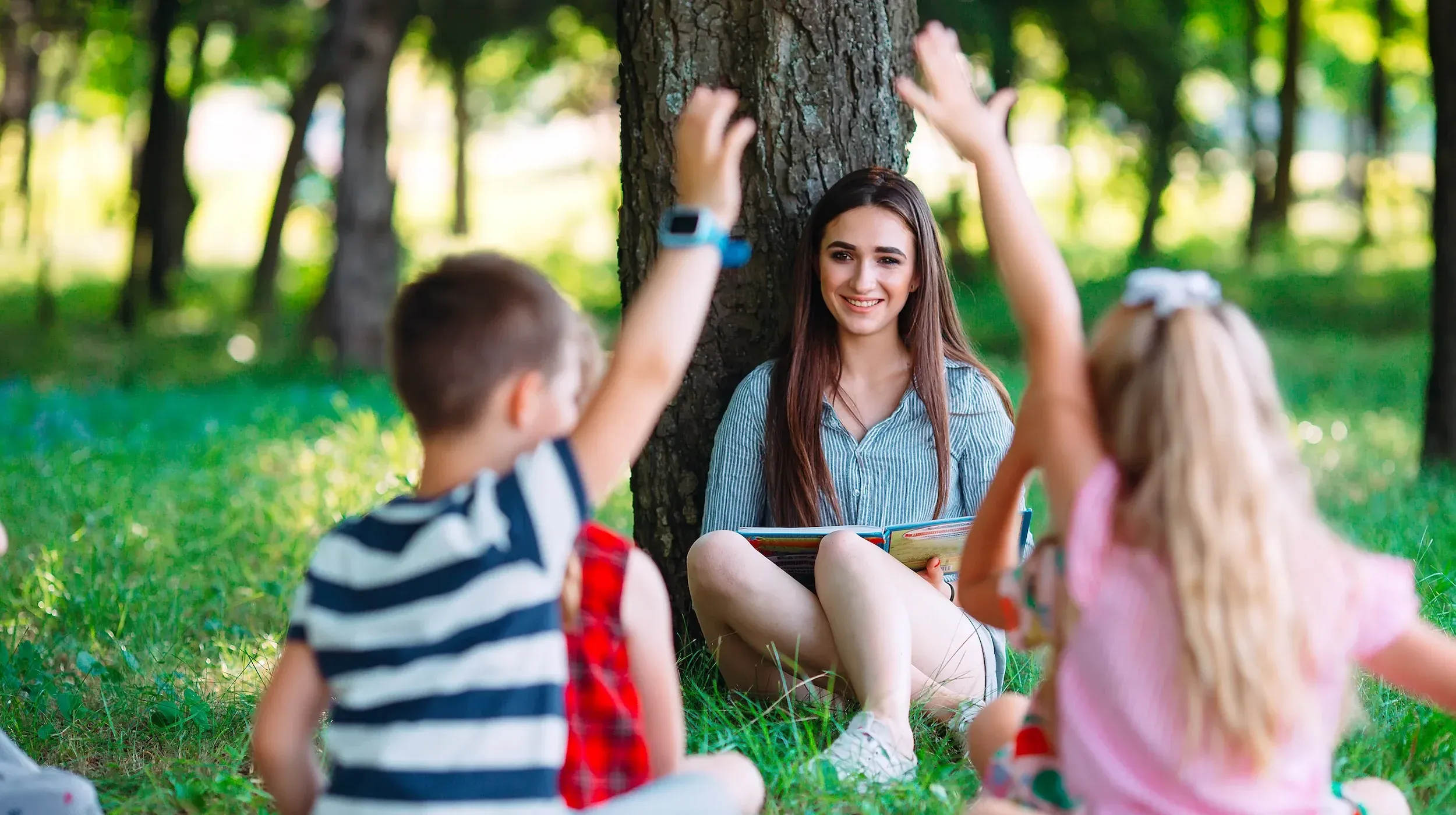 Foto di una classe che fa lezione sul prato. I bambini di spalle alzano la mano e la maestra sorride