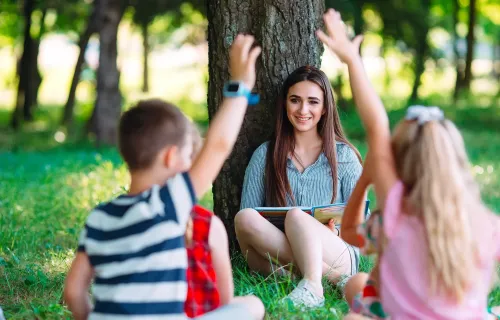 Foto di una classe che fa lezione sul prato. I bambini di spalle alzano la mano e la maestra sorride