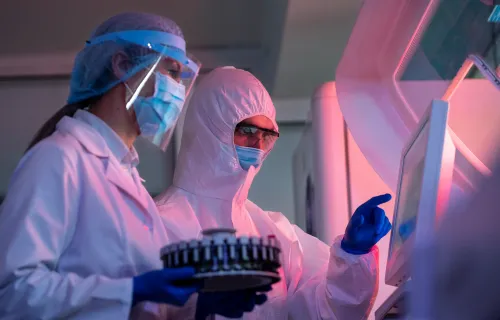 Foto di due ricercatori in un laboratorio con delle tute protettive e che lavorano con un macchinario